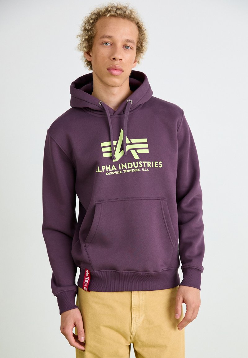Sweat-shirt à capuche violet avec poche kangourou à l'avant, mettant en avant un logo graphique jaune et une accent de tag rouge à l'ourlet. Tissu lisse, coupe décontractée.