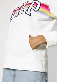 Felpa bianca con il logo "GAP" in lettere grasse e un motivo a strisce arcobaleno. Presenta una tasca frontale e polsini a costine. Tessuto morbido.