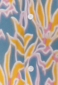 Stoff mit Blumenmuster in Gelb, Pink und Weiß auf blauem Hintergrund, mit einem weißen Knopf als auffälliges Detail.