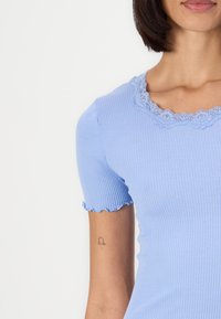 Vaaleansininen ribbed-paitatoppi, jossa on aaltoileva pitsikaulus. Ominaisuuksiin kuuluu istuva muotoilu ja hieman röyhelöity hihansuu.