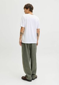 Witte korte mouwen t-shirt en losse olijfgroene broek. Materiaal heeft een zachte textuur. Bruine schoenen met zwarte zolen. Tattoeages zichtbaar op de armen.