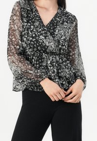 Schwarze transparente Bluse mit weißem Blumenmuster, V-Ausschnitt und langen Puffärmeln, tailliert für einen blusentypischen Effekt.