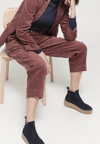 Terrakottabrunt kordflorsdräkt med en figursydd jacka och korta byxor, tillsammans med marinblå mocka ankelstövlar med texturerade detaljer.