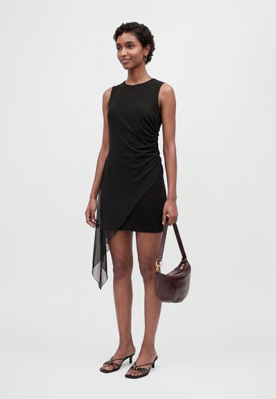 Une femme porte une robe noire asymétrique avec un pan drapé en tissu transparent, un sac à bandoulière bordeaux et des sandales à talons.