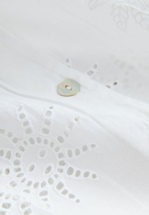 Krupni plan bijele tkanine ukrašene vezenim otvorima u obliku sunca i malim, okruglim, iridescentnim gumbom prišivenim na nju.