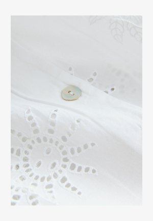 Közelkép fehér szövetről, amelyen nap alakú lyukacsos hímzés és egy kis, kerek, irizáló gomb van varrva.