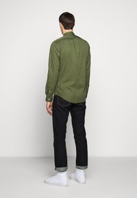 Snygg olivgrön skjorta med långa ärmar, knappad krage och rundad fåll, matchad med mörk denimjeans och vita höga sneakers.