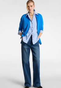 Cardigan blu chiaro sopra una camicia a righe bianche e blu con bottoni, abbinato a jeans a gamba larga blu scuro. Tessuto morbido con una vestibilità rilassata.