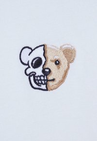 Geborduurd ontwerp met een helft schedel en een helft berengezicht in zwart en beige op een lichtgrijze achtergrond.