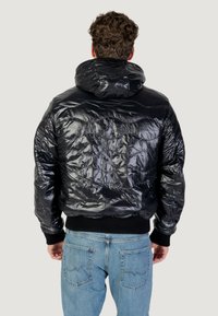 Giacca puffa nera con una texture lucida e trapuntata, dotata di cappuccio e polsini a costine. Abbinata a jeans in denim azzurro chiaro.