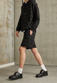 Haut à capuche noir et shorts avec des motifs de visages souriants jaunes. Porté avec des chaussettes blanches et des chaussures noires à lacets. Tissu lisse, coupe décontractée.