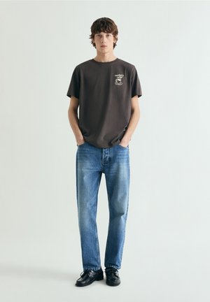 Tânăr stând cu mâinile în buzunare, purtând un tricou negru cu imprimeu grafic, blugi albaștri și pantofi negri, pe un fundal alb simplu.