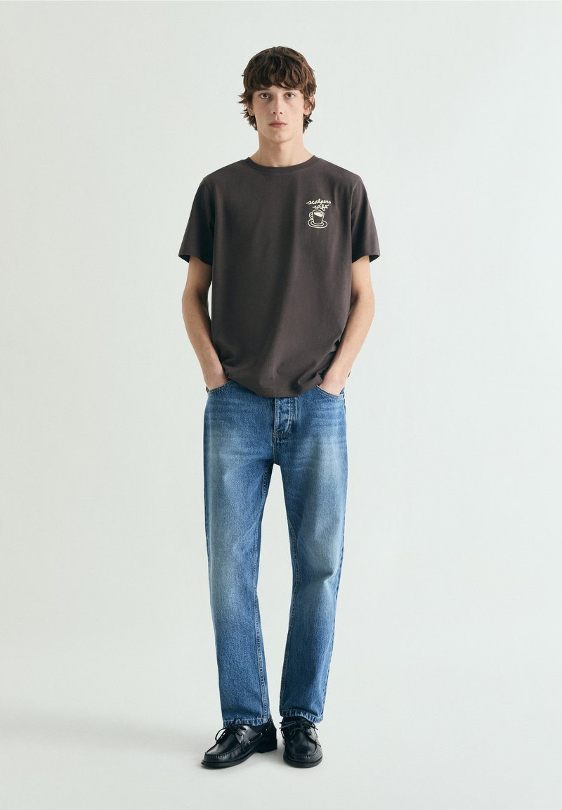 Ung mand stående med hænderne i lommerne iført sort t-shirt med grafik, blå jeans og sorte sko på ensfarvet hvid baggrund.