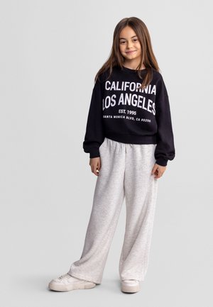 Jeune fille aux longs cheveux bruns portant un sweat-shirt noir avec le texte "California Los Angeles", un pantalon large gris clair et des baskets blanches.