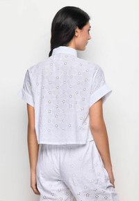 Camicia corta bianca in cotone con maniche corte, caratterizzata da motivi floreali ricamati e dettagli a occhiello, design leggero e arioso.