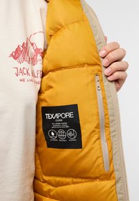 Intérieur d'une veste isolée jaune avec doublure beige. Comprend une poche zippée et une étiquette TEXAPORE indiquant des propriétés imperméables, respirantes et coupe-vent.