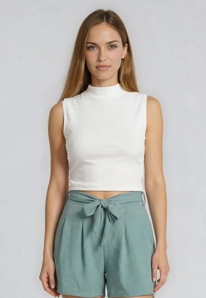 Jonge vrouw met lang lichtbruin haar, draagend een mouwloos wit topje en hooggetailleerde turquoise shorts met een strik aan de voorkant, staand tegen een effen achtergrond.