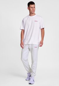 Weißes T-Shirt mit kurzen Ärmeln und kleinem Logo auf der Brust. Kombiniert mit hellgrauen Jogginghosen und Sportschuhen mit lila Akzenten.