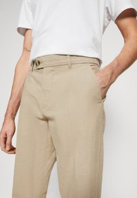 Pantalons en lin beige avec une coupe droite. Ils présentent une fermeture à crochet à l'avant et des poches latérales. Texture lisse et matériau léger.