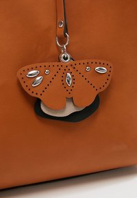 Pomarańczowa skórzana torba z brelokiem w kształcie motyla, ozdobionym srebrnymi ćwiekami i perforowanymi detalami.