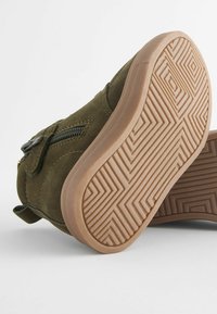 Groene suède schoen met een ritsluiting, een gestructureerde bruine rubberen zool met een zigzagpatroon en een gevoerde enkelsteun.