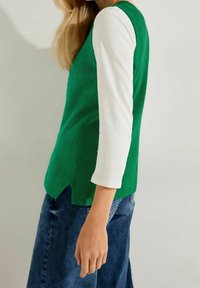 Débardeur côtelé vert avec une chemise blanche à manches longues en dessous, présentant un col en V et une fente latérale. Porté avec un jean denim bleu.