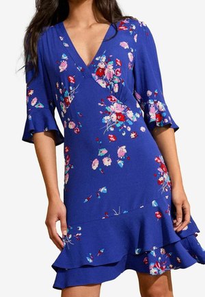 Vrouw draagt een blauw bloemen wikkeljurk met gerafelde mouwen en zoom, met rode, roze en lichtblauwe bloemenpatronen.