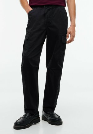 Ein Mann trägt schwarze Cargohosen und schwarze Lederloafers und steht mit einer Hand in der Tasche vor einem einfarbigen Hintergrund.