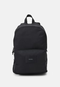 Czarny plecak wykonany z trwałej tkaniny, wyposażony w przednią kieszeń, zamknięcie na zamek oraz naszywkę z logo. Prosty design z regulowanymi paskami.