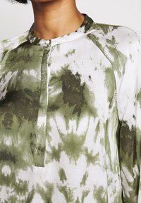 Koszula z długim rękawem w białym i zielonym wzorze tie-dye, z zapięciem na guziki, kołnierzem mandaryńskim i rękawami raglanowymi. Gładka tekstura materiału.