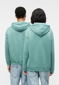 Φούτερ με κουκούλα σε απαλό μέντα πράσινο, με ριμπ μανσέτες και τελειώματα, εμφανίζονται από πίσω με χαλαρή εφαρμογή. Συνδυασμένα με ανοιχτό τζιν.