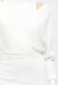 Frau trägt ein weißes, geripptes Strickkleid mit V-Ausschnitt und Cut-out-Details an den Schultern, das die strukturierte Stoffstruktur und die taillierte Passform zeigt.