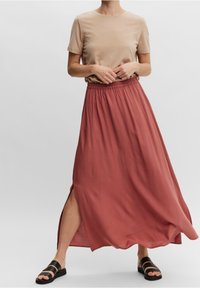 Longue jupe maxi couleur rouille avec une taille élastique et une fente latérale, associée à un t-shirt beige ajusté et des sandales noires à lanières.