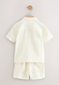 Cămașă cu mâneci scurte și pantaloni scurți ivoar, asortați, realizați dintr-un material texturat cu modele în relief, atârnați pe un umeraș de lemn pe un fundal simplu.