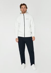 Sudadera blanca con cremallera y capucha ajustable, bolsillos frontales y cremallera negra; combinada con pantalones azul marino y zapatillas blancas.