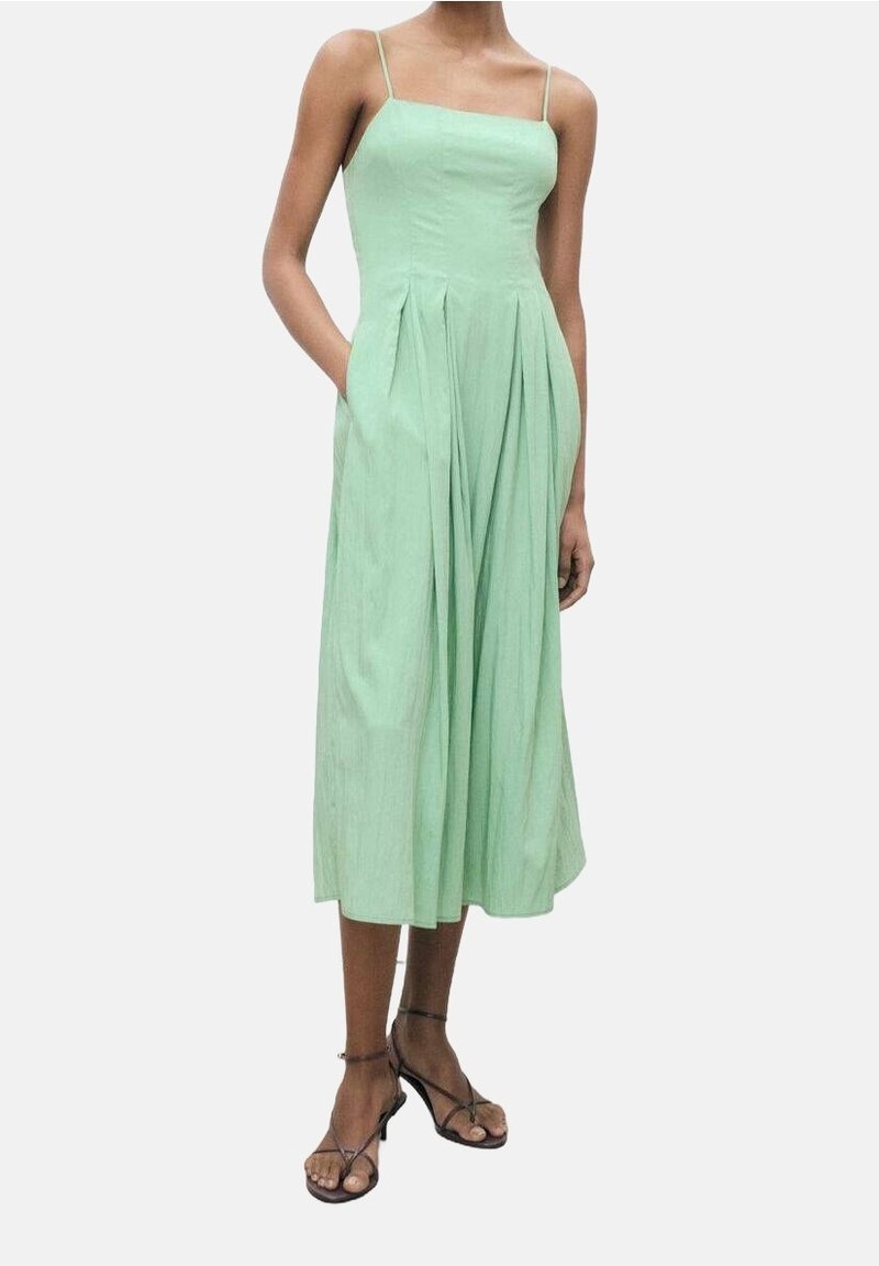Femme portant une robe midi sans manches, vert clair, avec de fines bretelles et des plis, associée à des sandales noires à talons bas et fines brides, debout avec une main dans la poche.