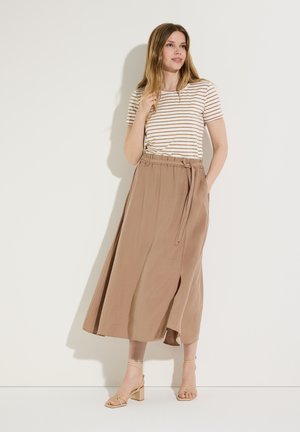 Frau trägt beige gestreiftes Kurzarmshirt, hoch taillierten braunen Midirock mit seitlichem Schlitz und beige Sandalen mit Blockabsatz, steht vor weißer Wand.