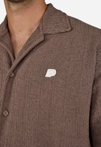 Barna, texturált ing, amelynek pöttyös gallérja van, és egy kis fehér hímzett 'P' logóval, valamint gombos záródással rendelkezik. A bordázott anyag diszkrét részletet ad az inghez.