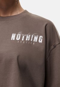 Felpa di cotone marrone con collo rotondo, caratterizzata da un testo grafico in rilievo bianco "Goods For NOTHING ATELIER" sul fronte.
