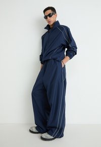 Navy tracksuit with a high collar, featuring gray piping details. Loose-fitting pants and oversized top, paired with gray slip-on shoes.