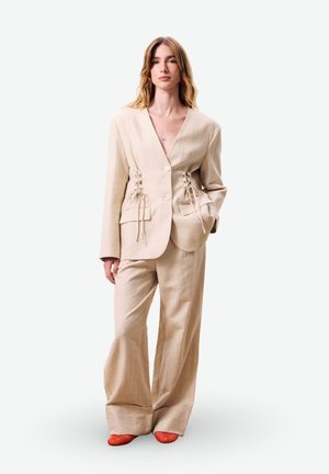 Vrouw in een beige oversized pak met veterdetail in de taille en broek met wijde pijpen, gecombineerd met rode textuurschoenen, staand tegen een witte achtergrond.