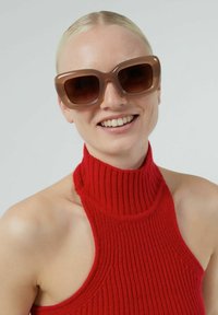 Bruna överdimensionerade solglasögon med en glansig finish och fyrkantiga bågar. Bärs tillsammans med en röd ribbad topp med hög hals. Slät textur, minimal hårdvara.