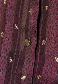 Blusa trasparente viola con motivi neri diagonali e dettagli dorati. Presenta un bottone nero rotondo al centro, che mette in evidenza il suo design elegante.