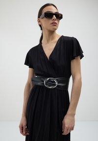 Donna che indossa un vestito avvolgente nero con maniche a sbuffo, una larga cintura nera con una grande fibbia d'argento e occhiali da sole neri rettangolari, su uno sfondo bianco.