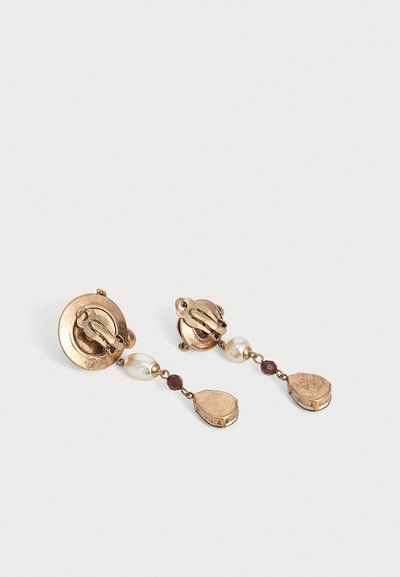 Paire de boucles d'oreilles clips vintage dorées avec des fausses perles pendantes et de petites perles sombres sur un fond clair uni.