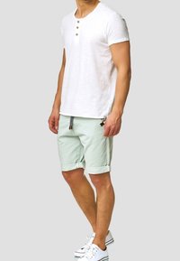 Camiseta blanca de manga corta con tres botones de madera. Pantalones cortos verde claro, doblados en el dobladillo, con cordón y bolsillos laterales. Zapatillas blancas.