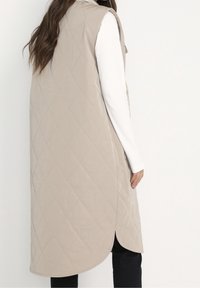 Beige gesteppte Weste mit hohem Kragen und Seitenschlitzen, kombiniert mit einem weißen Langarmshirt und schwarzen Hosen. Glatte Textur und minimalistisches Design.