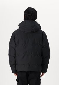 Zwarte gewatteerde jas met capuchon, voorzien van een gequilt ontwerp en een zachte textuur. Achteraan is er een ontspannen pasvorm te zien met minimale details.