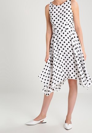 Kvinde iført en hvid ærmeløs kjole med sorte polkaprikker og en asymmetrisk skørtkant, kombineret med hvide slingback-flats.