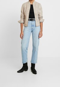 Beige bomber bunda s žebrovanými detaily, černé tričko, světle modré džíny rovného střihu a černé kotníkové boty. Hladká textura, moderní střih.